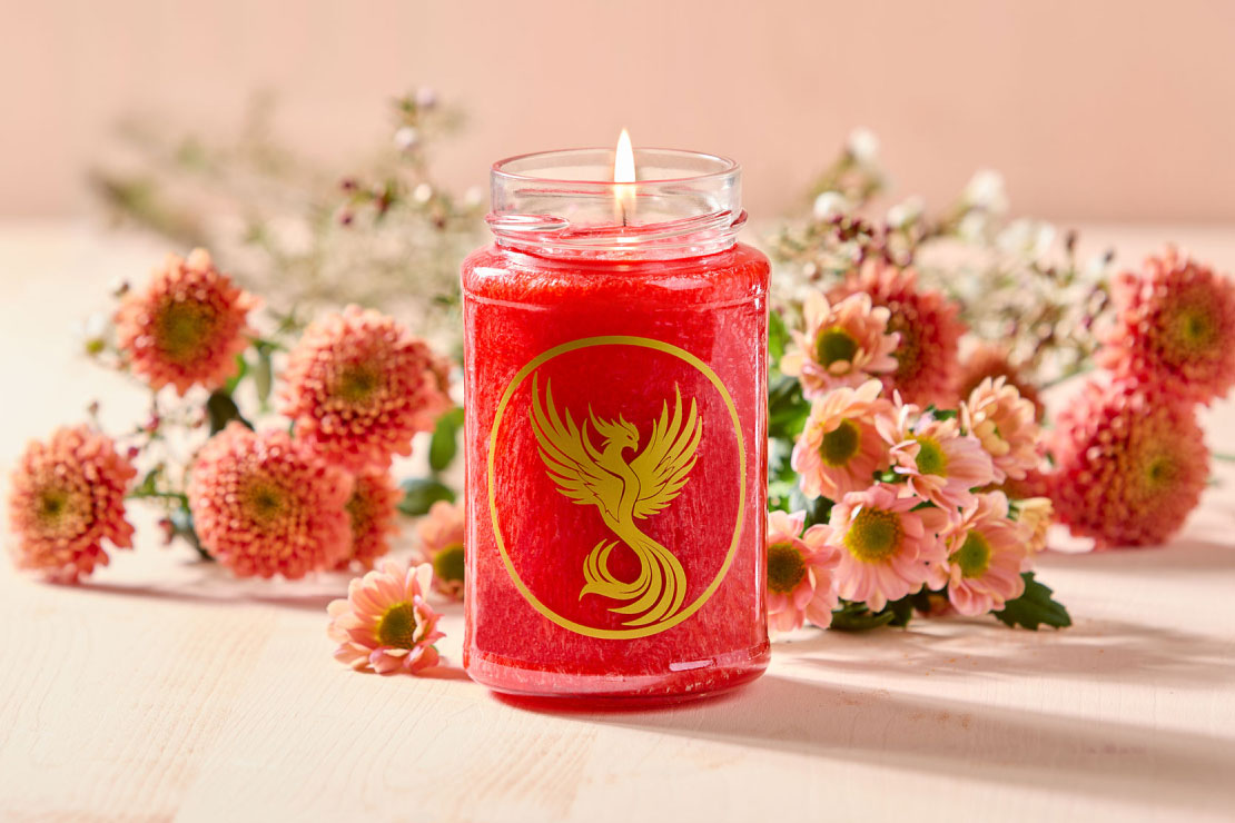 Eine brennende rote Duftkerze im Glas mit aufgedrucktem goldenen Phönix-Emblem auf der Vorderseite. Um die Kerze herum sind zarte Blüten arrangiert. 
