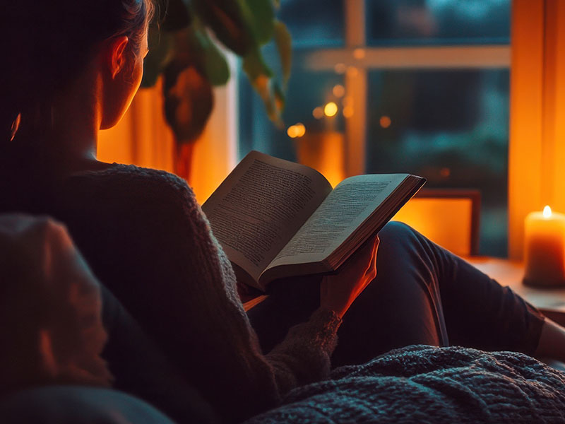 Frau liest abends ein Buch bei Kerzenlicht am Fenster. Draußen ist es dunkel, drinnen herrscht eine warme, behagliche Atmosphäre – ein Moment der Stille und Geborgenheit.
