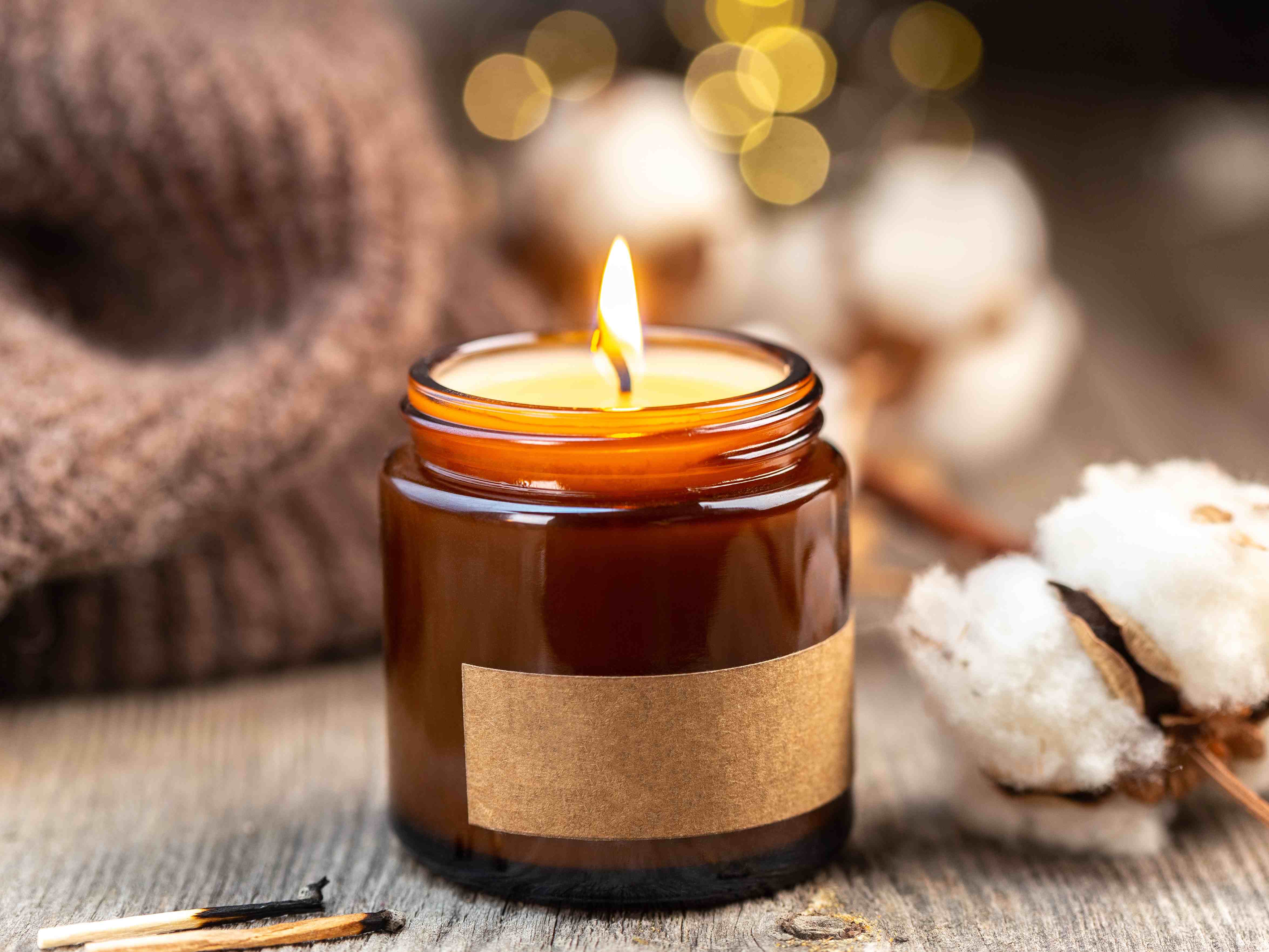 Brennende Kerze in einem braunen Glas auf Holztisch, daneben Baumwollzweige und ein gestrickter Schal vor warmen Lichtpunkten im Hintergrund.