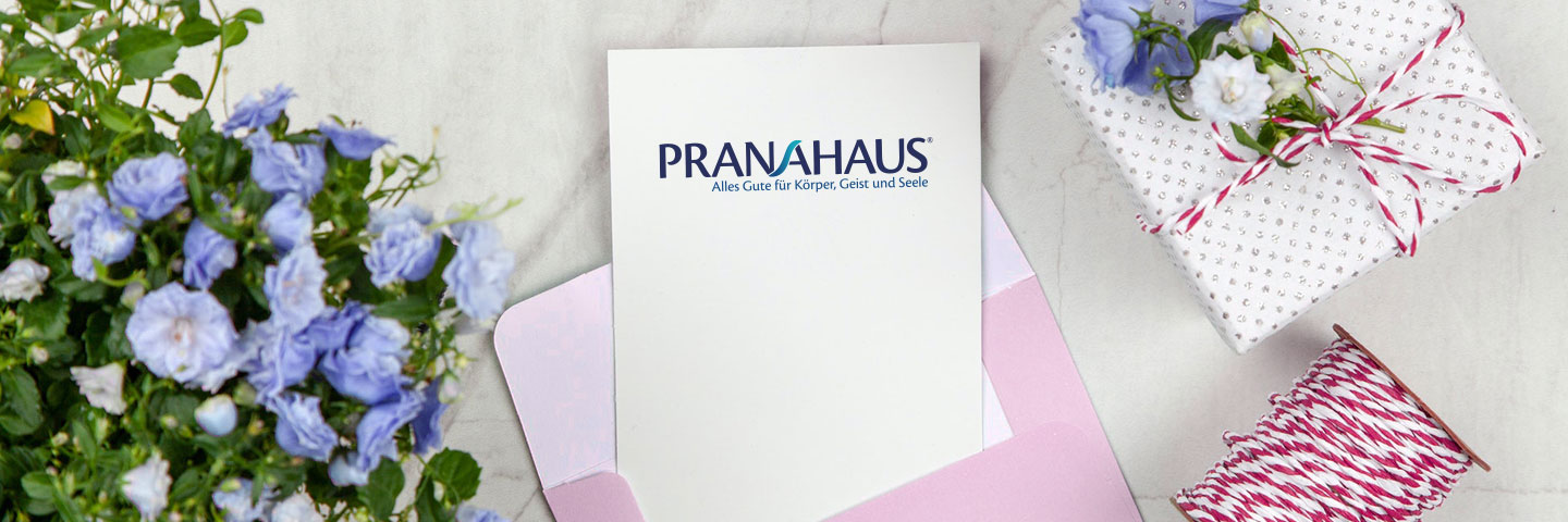 Karte mit PranaHaus-Logo in einem Umschlag, daneben ein Geschenk und ein Blumenstrauß