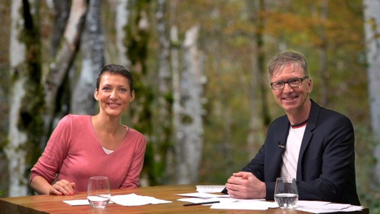 Ina Rudolph und der Moderator des Webinars sitzen an einem Tisch und lächeln in die Kamera