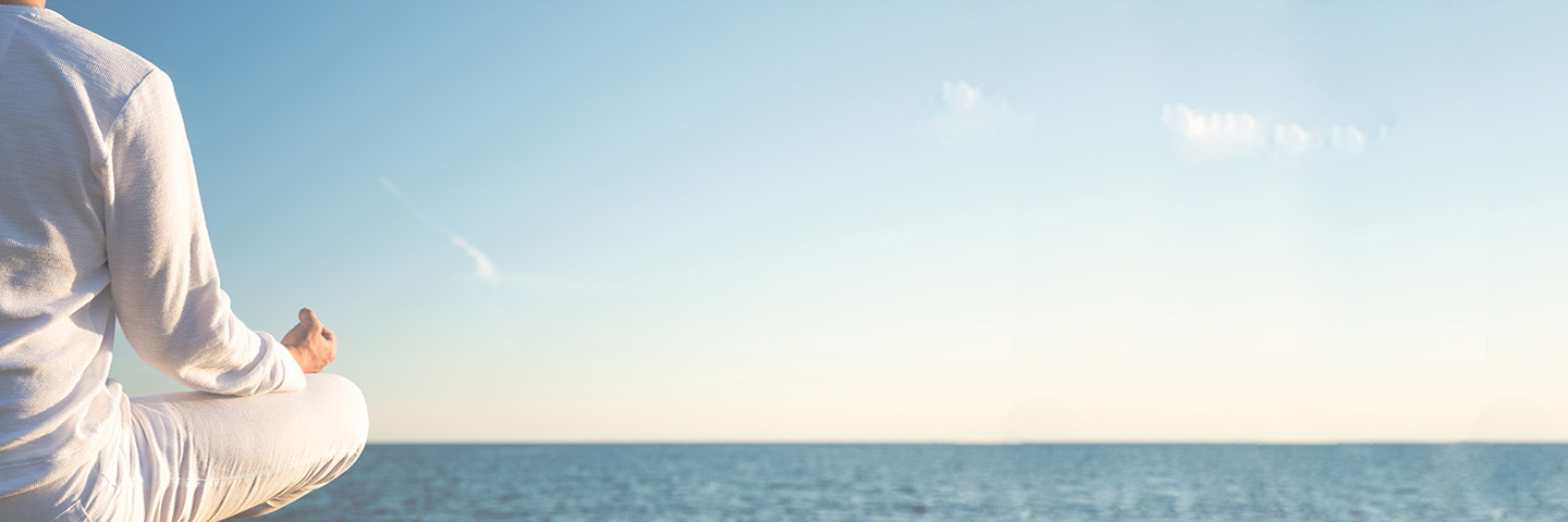 Mann in weißer Kleidung sitzt mit dem Rücken zur Kamera am Meer und meditiert