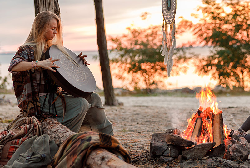 Eine Frau sitzt an einem Lagerfeuer und hält eine Trommel in der Hand.