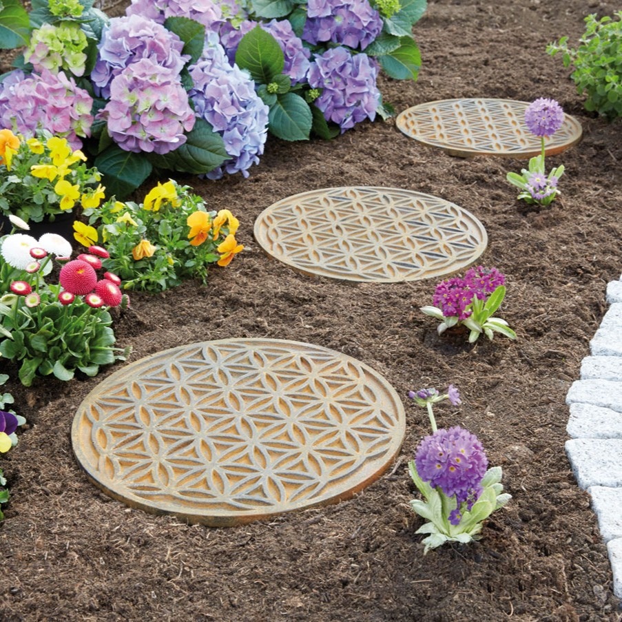 Drei runde Platten mit ornamentalem Muster liegen in einem frisch bepflanzten Blumenbeet mit lila, gelben und pinken Blüten.