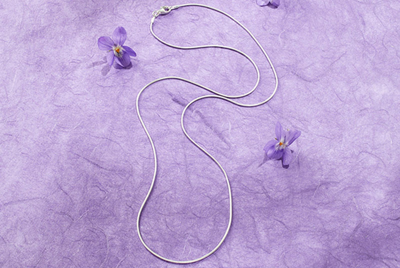 Eine schlichte Silberkette liegt auf einem violetten Untergrund, daneben violette Veilchenblüten.