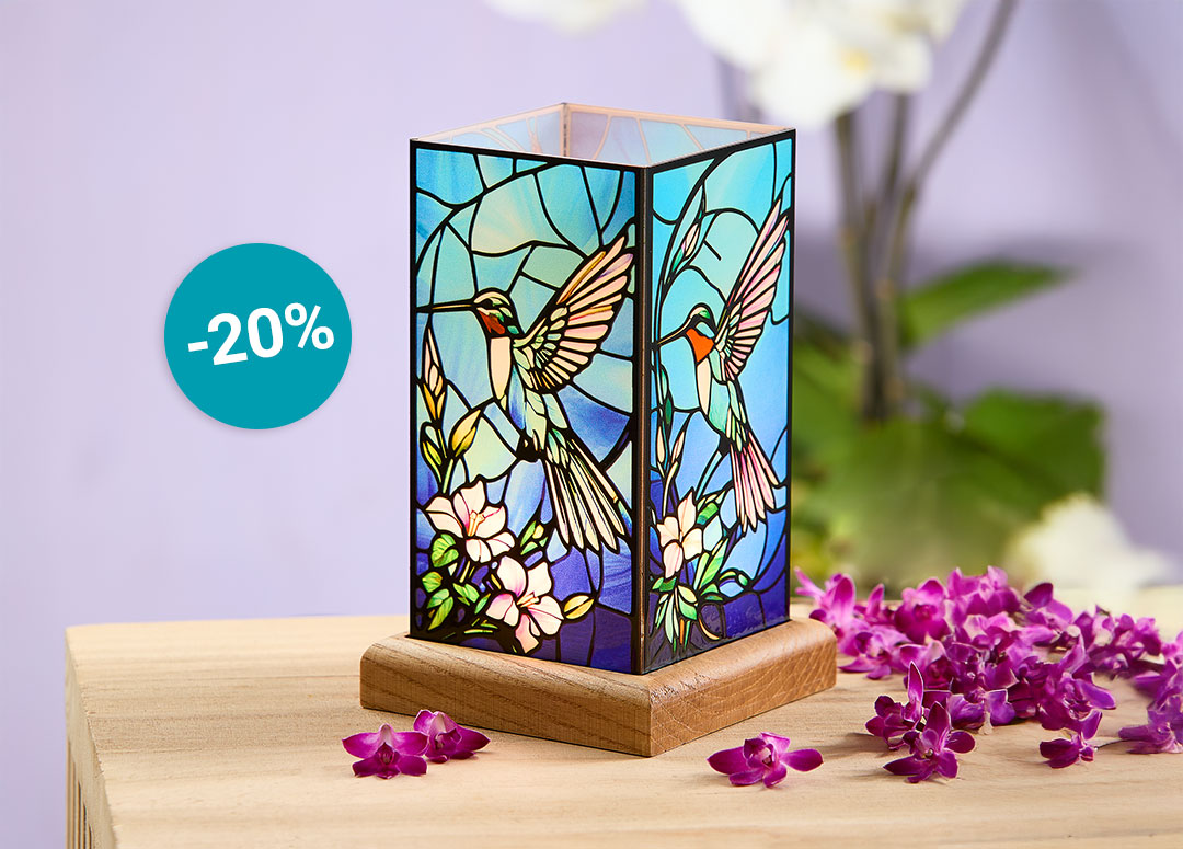 Quadratische Tischlampe im Tiffany-Stil mit farbigem Glasmotiv aus Kolibris und Blüten auf Holzsockel. Daneben liegen violette Blüten, links ein Hinweis „-20 %“. Die Szene wirkt dekorativ, lichtvoll und bringt Farbe ins zu Hause.
