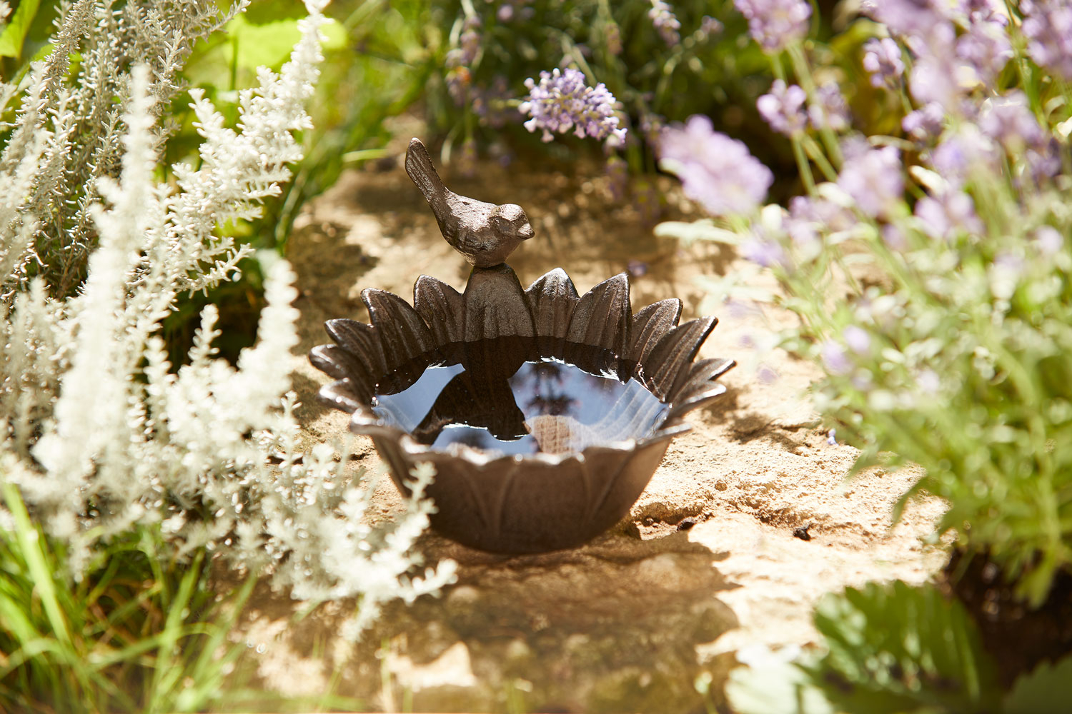 Eine dekorative Vogeltränke aus Gusseisen in Form einer Sonnenblume steht auf einem sonnigen Gartenweg aus Natursteinen. Auf dem Rand der Tränke sitzt eine kleine Metallfigur in Form eines Vogels.