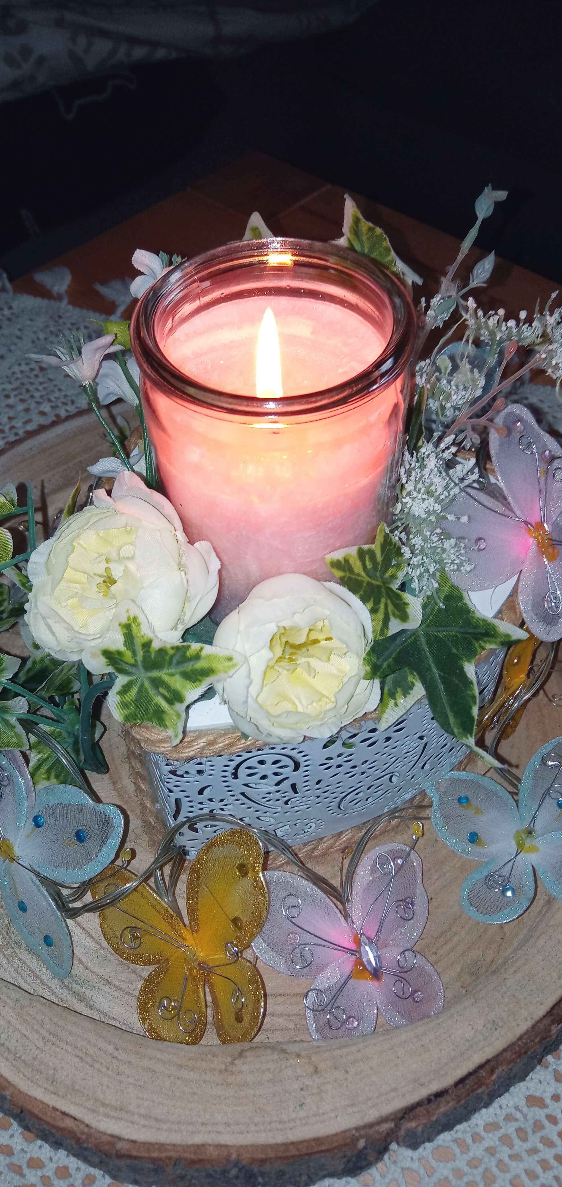 Brennende Kerze auf einer Holzscheibe, geschmückt mit Blumen und einer Kette mit bunten Schmetterlingen