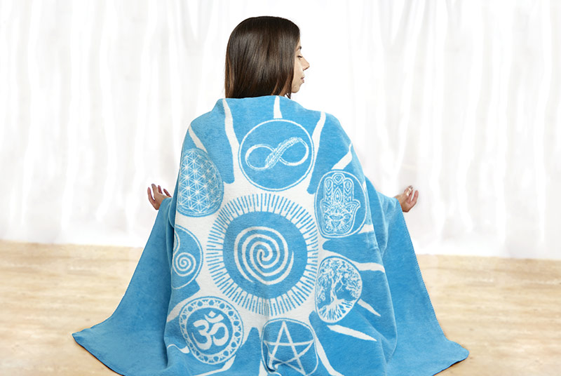 Eine Frau sitzt in Meditationshaltung auf dem Boden und hat eine Meditationsdecke um sich geschlungen. Die Decke ist türkisblaue mit aufgedruckten weißen Energiesymbolen.