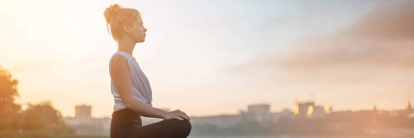 Eine junge Frau sitzt im Schneidersitz und hat die Hände auf den Knien abgelegt. Sie meditiert, während im Hintergrund die Sonne aufgeht.