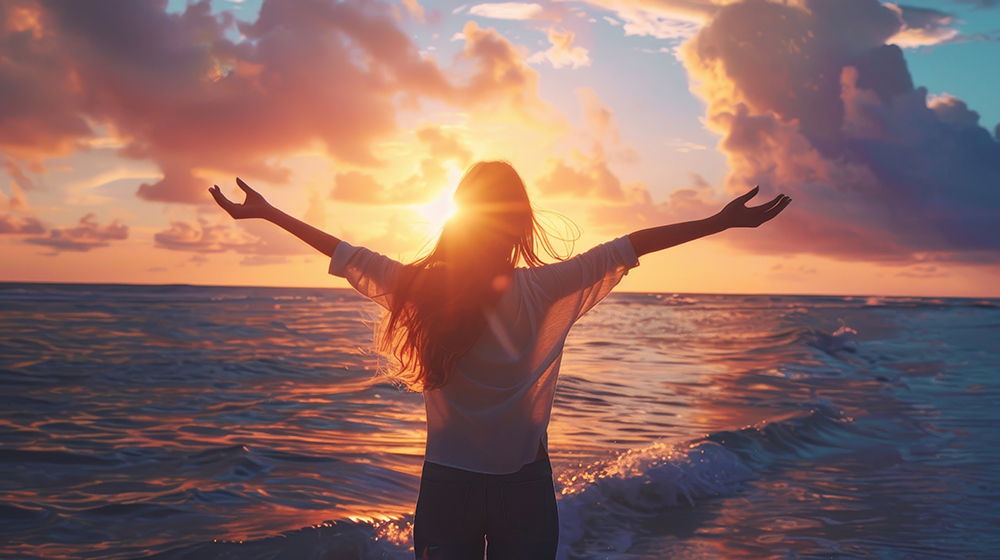 Eine Frau steht mit ausgebreiteten Armen am Meer und blickt in einen leuchtenden Sonnenuntergang, der den Himmel und das Wasser in warme Farben taucht.