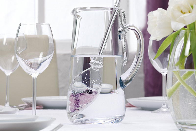 Auf einem gedeckten Tisch steht eine Glaskaraffe, darin ein Vita-Juwel-Stab mit Edelsteinen. Rechts daneben steht eine Vase mit weißen Tulpen.