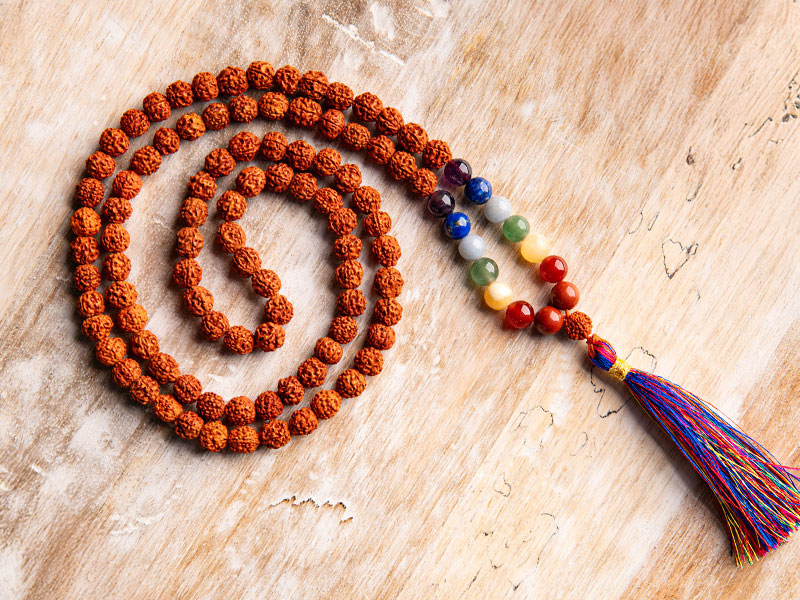 Spiralförmig gelegte Mala aus braunen Rudraksha-Perlen mit farbigen Edelsteinen für die sieben Chakren und einer bunten Quaste, auf hellem Holzuntergrund – ein harmonisches Symbol spiritueller Ausrichtung.