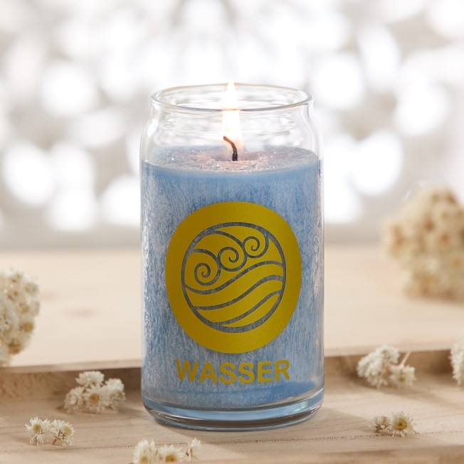 Brennende blaue Kerze im Glas mit der Aufschrift „WASSER“ auf einem hellen Holztisch, umgeben von kleinen Trockenblumen.