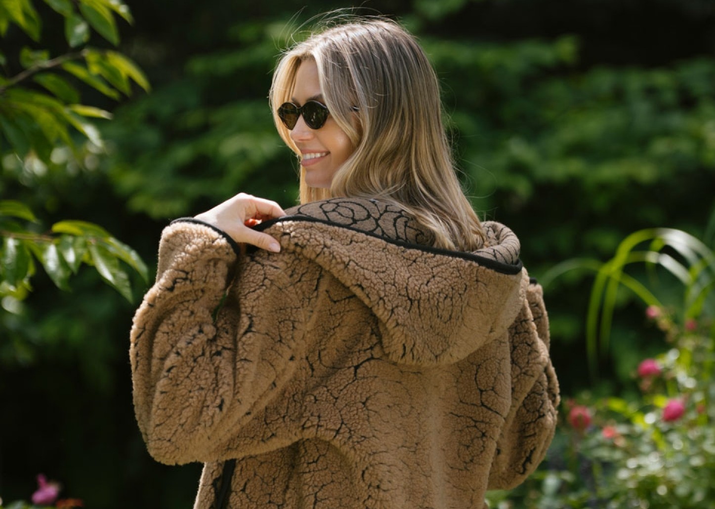 Eine lächelnde Frau mit langen blonden Haaren und Sonnenbrille trägt eine braune Strickjacke mit Kapuze und steht in einem grünen Garten.