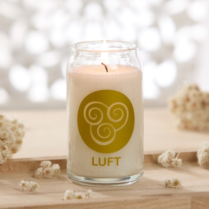 Brennende weiße Kerze im Glas mit der Aufschrift „LUFT“ auf einem hellen Holztisch, umgeben von kleinen Trockenblumen.