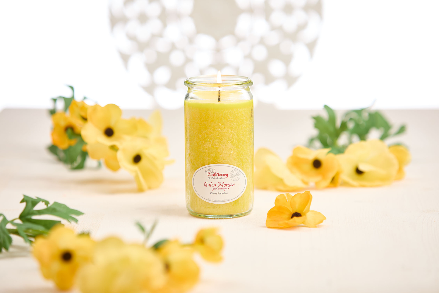 Gelbe Duftkerze mit Etikett „Guten Morgen“, auf hellem Tisch mit gelben Blüten dekoriert. Die helle, freundliche Szenerie weckt Leichtigkeit und Frische für einen beschwingten Tagesbeginn.