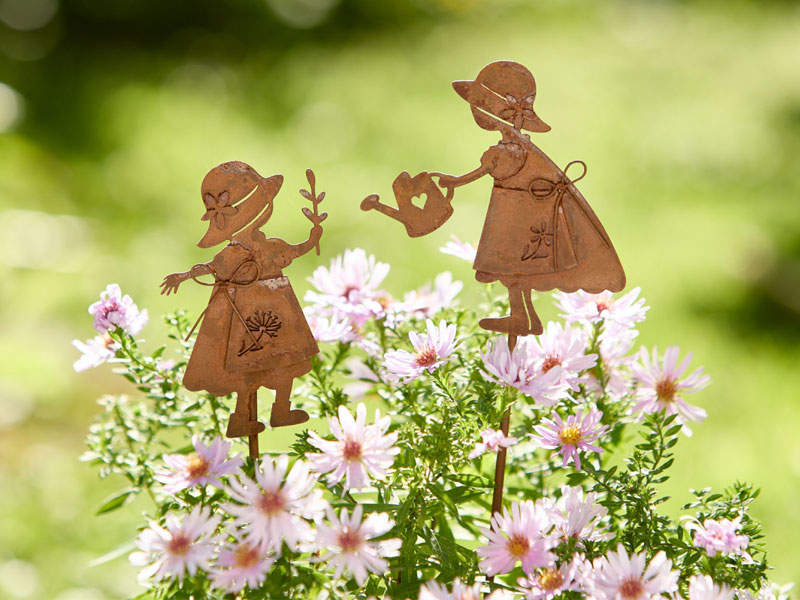 Zwei rostfarbene Metallfiguren in Form von Mädchen mit Gießkanne stehen als Gartenstecker zwischen zarten rosa Blüten. Die Szene wirkt verspielt, frühlingshaft und bringt liebevolle Leichtigkeit ins Beet.
