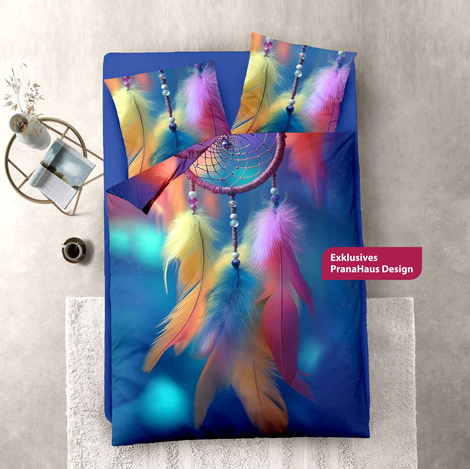 Bunte Bettwäsche mit einem großflächigen Traumfänger-Motiv aus farbigen Federn und Perlen, auf einem gemachten Bett mit Beistelltisch, Tasse und Deko auf hellem Boden.