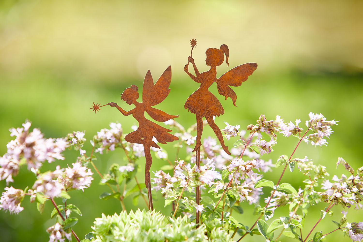 Zwei Edelrost-Gartenstecker in Form von Feen stehen zwischen blühenden Kräutern. Beide Feen haben feine Flügel und halten einen Zauberstab mit sternförmiger Spitze.
