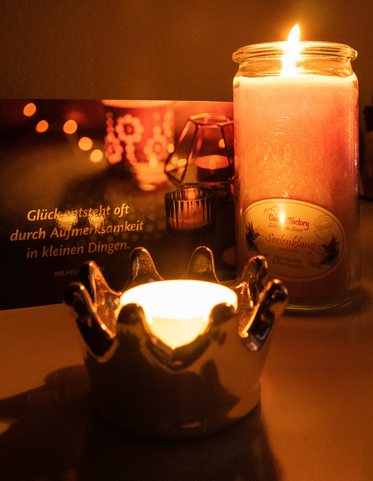 Brennende Kerze und ein Teelicht in einem kronenförmigen Halter neben einer Postkarte, auf der ebenfalls Kerzen abgebildet sind sowie das Zitat "Glück entsteht oft durch Aufmerksamkeit in kleinen Dingen." von Wilhelm Busch