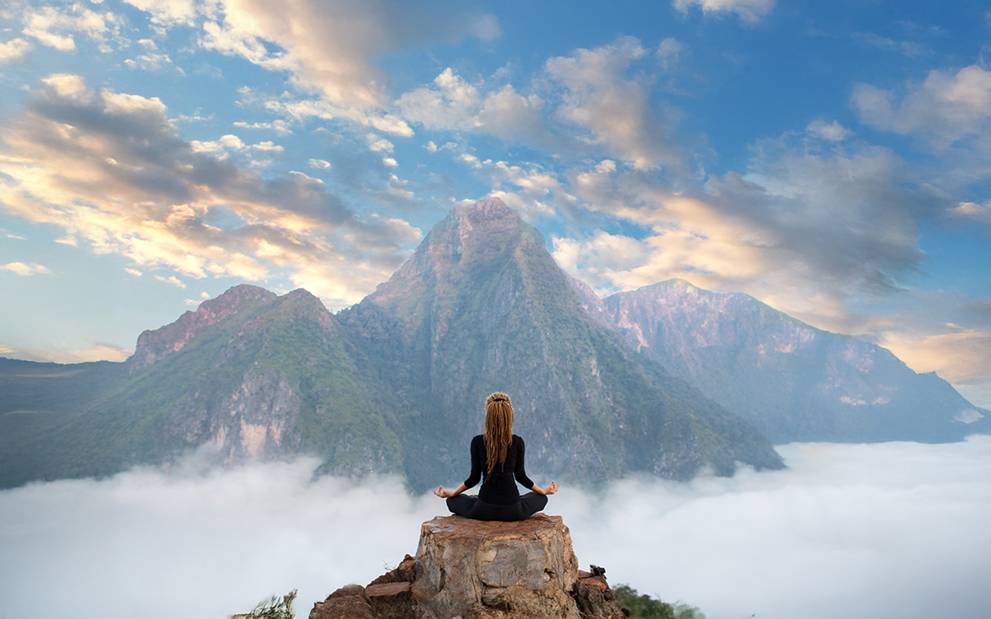 Eine Frau meditiert im Lotussitz auf einem Felsen über den Wolken mit Blick auf ein majestätisches, von Nebel umhülltes Bergpanorama unter einem leicht bewölkten Himmel.