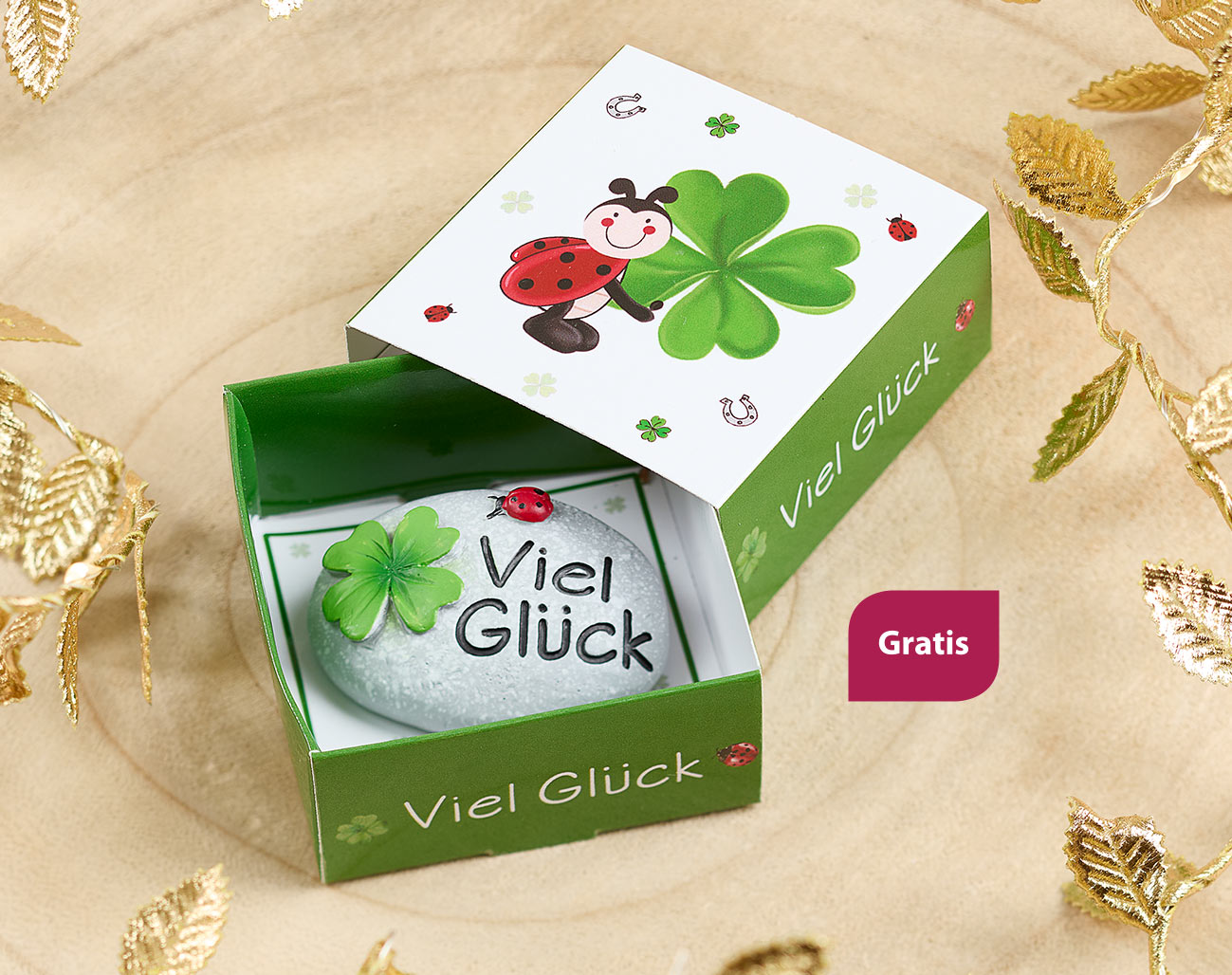 Geschenkverpackung mit Glücksmotiv, darin ein bemalter Stein mit Kleeblatt, Marienkäfer und der Aufschrift „Viel Glück“. Die grüne Box mit fröhlichem Design liegt auf hellem Holz mit goldenen Deko-Blättern.