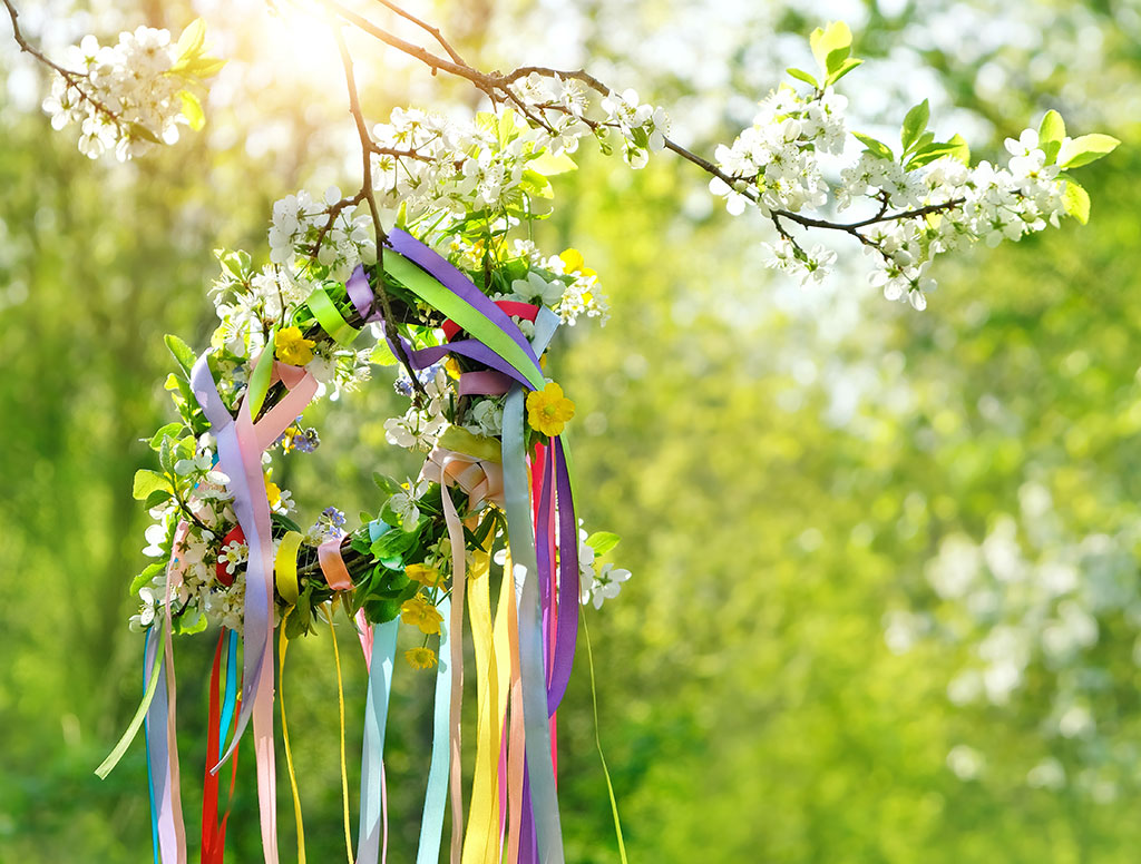 Bunter Frühlingskranz mit Blumen und langen, farbigen Bändern hängt an einem blühenden Zweig im Sonnenlicht vor einem grünen, weichen Hintergrund.