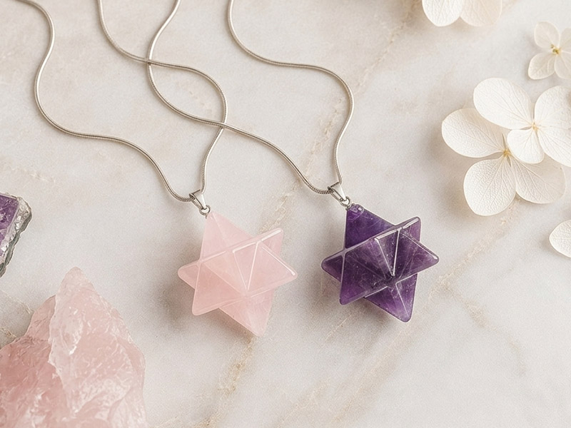 Zwei Edelstein-Anhänger in Merkaba-Form aus Rosenquarz und Amethyst an silberfarbenen Ketten auf heller Steinoberfläche mit Blüten und Kristallen.