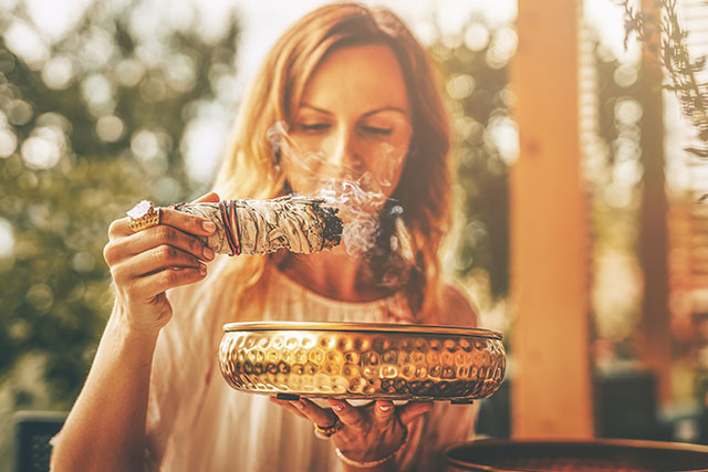 Eine Frau hält eine brennende Salbeibündel in der Hand, während sie eine goldene Schale darunter hält. Rauch steigt sanft auf, während das Licht einer sonnigen Umgebung die Szene durchflutet.