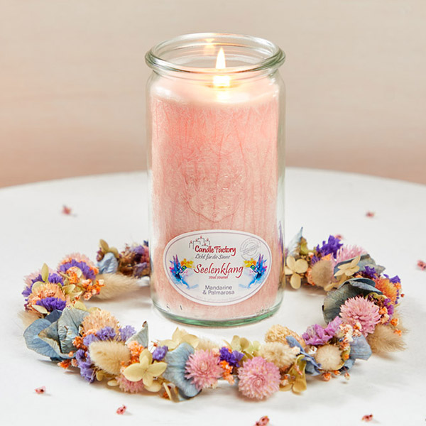 Eine brennende rosafarbene Duftkerze im Glas steht auf einem Tisch. Um die Kerze herum liegt ein dekorativer Kranz aus Trockenblumen in Pastellfarben, darunter Blau, Lila, Rosa und Beige. Kleine Blütenblätter sind auf dem Tisch verstreut.