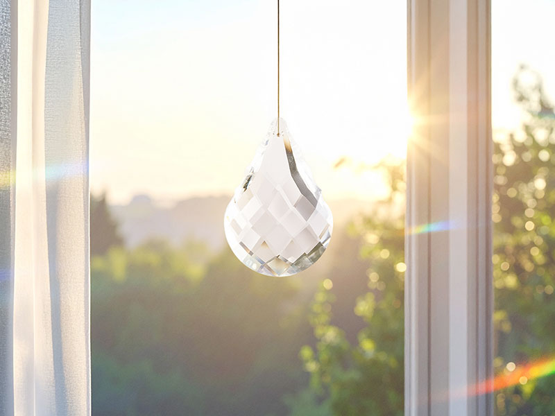 Tropfenförmiger Kristall-Sonnenfänger hängt am Fenster und fängt das einfallende Morgenlicht ein. Sanfte Regenbogenreflexe tanzen im Raum, draußen zeigt sich eine ruhige, grüne Landschaft im Sonnenaufgang.