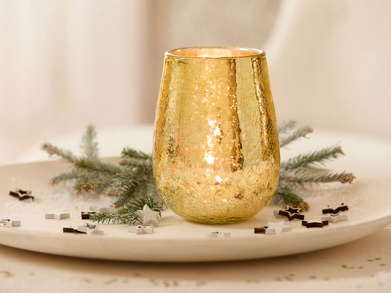 Ein glänzendes Windlicht in edler Goldoptik steht auf einem hellen Teller, umgeben von kleinen Tannenzweigen und winterlicher Streudeko in Sternform.